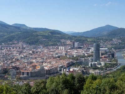 A panoramic view of the city of Bilbao, with a skyscraper and the shiny, pointy Guggenheim museum in the foreground. There's a river running though the city and mountains in the background.